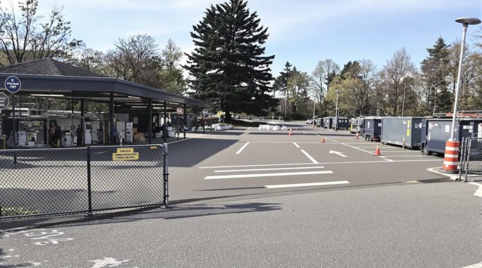 Brucker Blatt - Das Bild zeigt einen Außenbereich mit einem überdachten Gebäude auf der linken Seite. Vor dem Gebäude befindet sich ein Parkplatz mit weißen Markierungen und Pfeilen. Mehrere orangefarbene Verkehrskegel sind auf dem Parkplatz platziert. Hinter dem Parkplatz stehen mehrere graue Anhänger in einer Reihe. Im Hintergrund befinden sich Bäume und ein blauer Himmel mit einigen Wolken. Eine schwarze Kette mit einem gelben Schild ist im Vordergrund zu sehen.