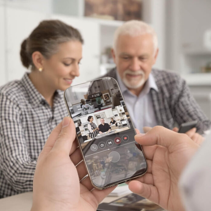 Brucker Blatt - Das Bild zeigt eine Nahaufnahme der Hände einer Person, die ein Smartphone hält, während im Hintergrund zwei verschwommene Personen, eine Frau und ein älterer Mann, zu sehen sind. Auf dem Smartphone-Bildschirm ist eine Videokonferenz mit mehreren Teilnehmern zu sehen. Die Frau im Hintergrund hat ihre Haare zu einem Knoten gebunden und trägt ein kariertes Hemd. Der Mann hat graue Haare und trägt ein kariertes Hemd über einem hellblauen Hemd. Beide scheinen auf den Bildschirm des Telefons zu schauen. Die Person, die das Telefon hält, scheint in einem Videoanruf aktiv zu sein, wie die Symbole auf dem Bildschirm andeuten, darunter Optionen für Mikrofonsteuerung und Beenden des Anrufs. Der Fokus liegt auf dem Smartphone und den Händen, wodurch der Hintergrund unscharf und weniger detailliert ist.