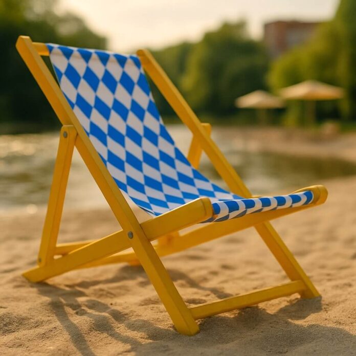 Brucker Blatt - Das Bild zeigt einen gelben Holzstrandkorb mit einem blau-weiß karierten Stoffbezug auf einer Sandfläche. Im Hintergrund sind ein Gewässer, grüne Vegetation und Gebäude erkennbar. Die Beleuchtung deutet auf Tageslicht hin.